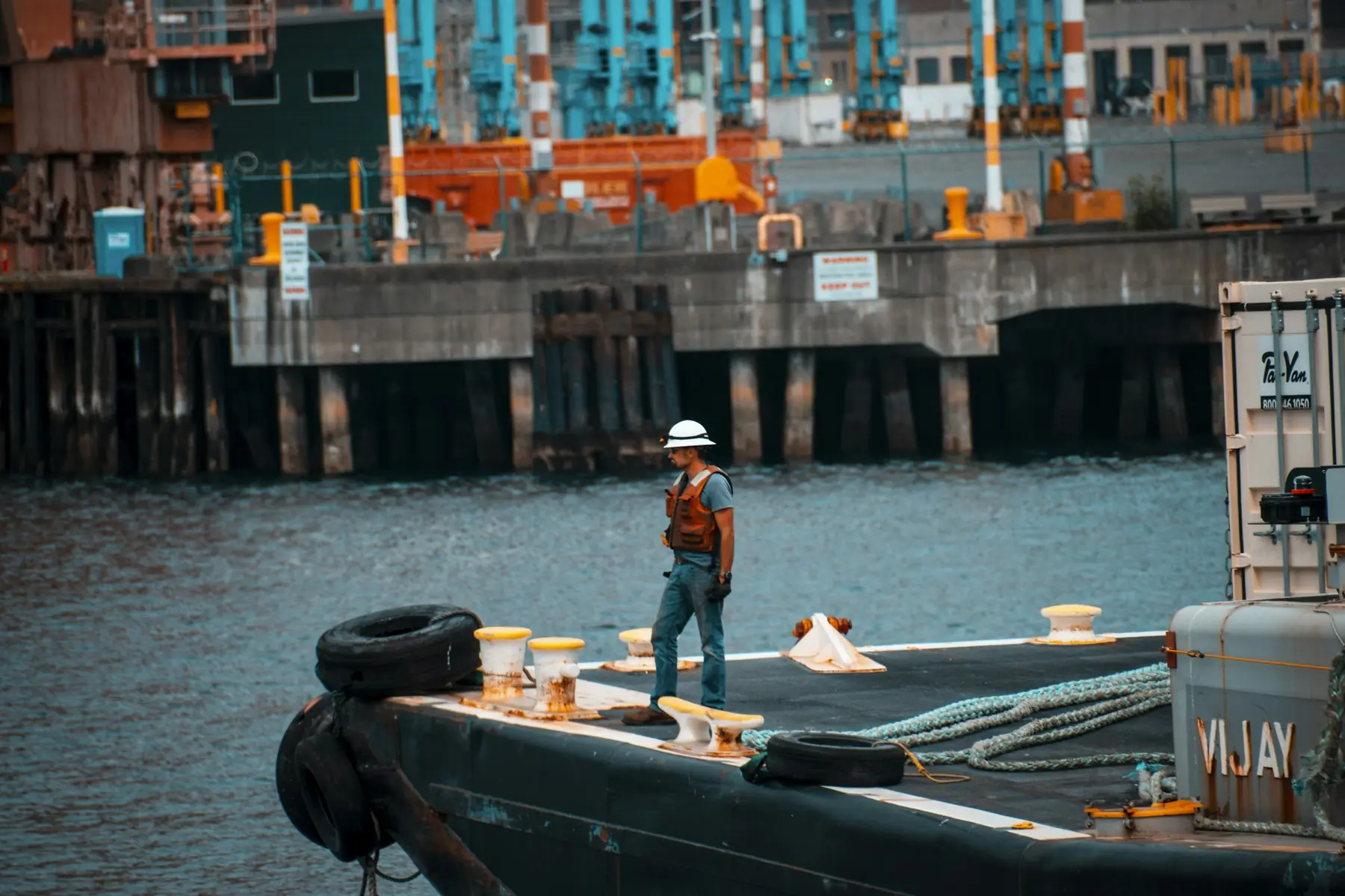 A deckhand in a hard hat and life vest working on a barge deck beside the pier