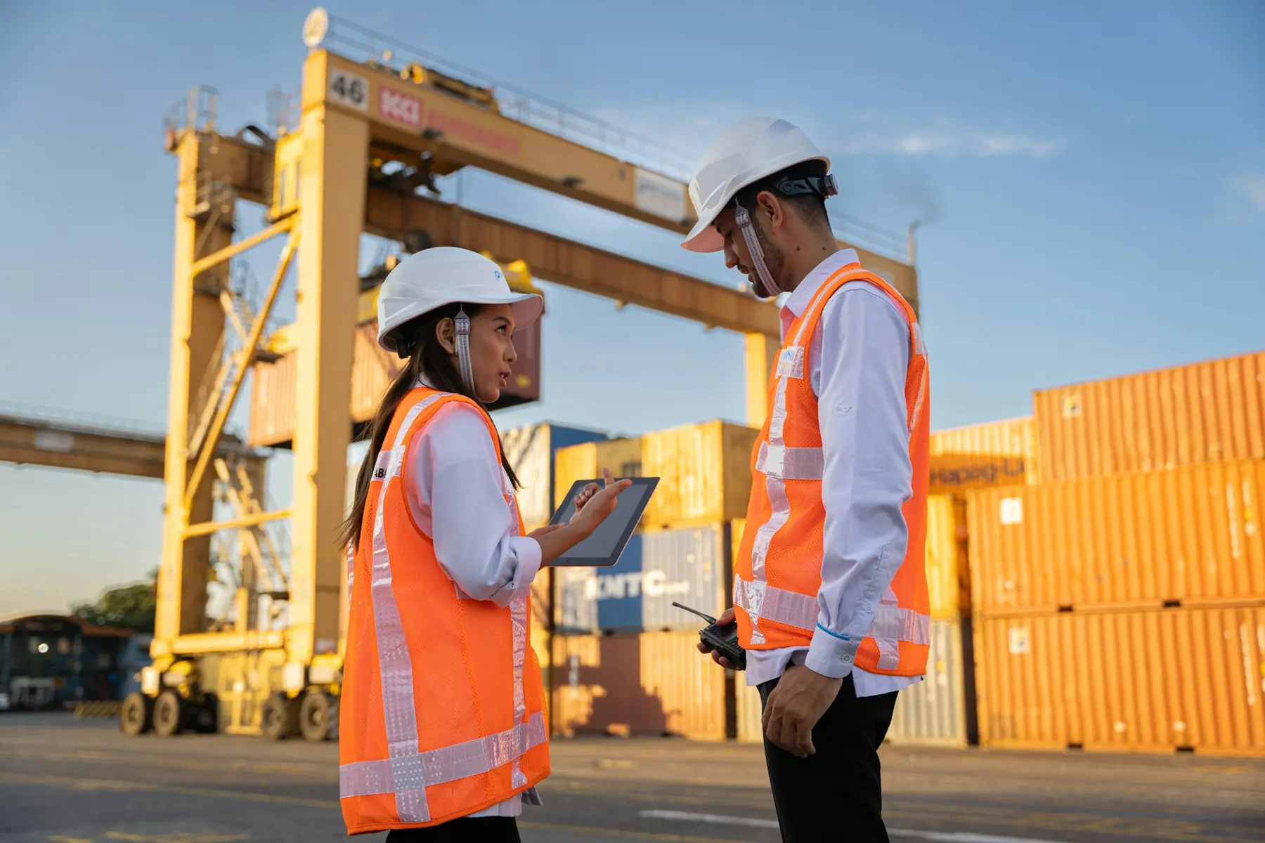 Two terminal workers in hard hats and vests reviewing a tablet beside a straddle carrier